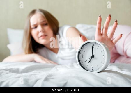 Verschlafen junge Frau wach in Schlafanzug schaltet Wecker in Eile. Morgendliche Routine und wachen Sie spät vom Wecker im Bett auf. Frau ist spät in Panik Stockfoto