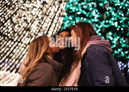 Weibliche Freunde küssen junge Frau auf den Wangen von Weihnachten Leuchtet Stockfoto