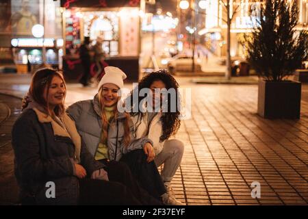 Glückliche Freundinnen sitzen auf der Straße in der Stadt in der Nacht Stockfoto