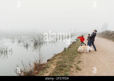 Vater und zwei Söhne mit Hund, die Steine in den See werfen An nebligen Tagen Stockfoto