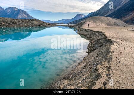 Wanderer, die an einem See durch ein entlegenes arktisches Tal an einem teilweise bewölkten Sommertag spazieren. Dramatische arktische Landschaft des Akshayuk Pass, Baffin Island, Kanada. Stockfoto