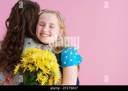 Happy stilvolle Mutter und Tochter mit langen welligen Haar mit gelben Chrysanthemen Blumen umarmen vor rosa Hintergrund. Stockfoto