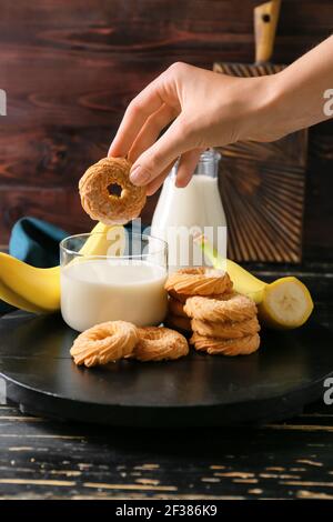 Weibliche Hand mit leckeren Bananenkeksen und Milch im Dunkeln Holzhintergrund Stockfoto