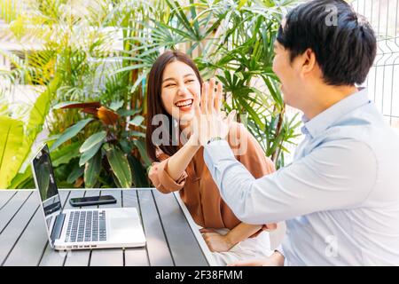 Fröhlich und aufgeregt junge asiatische Mann und Frau feiern gut nachrichten und Erfolg, indem sie einander hohe fünf geben Während Sie im modernen Café sitzen Stockfoto