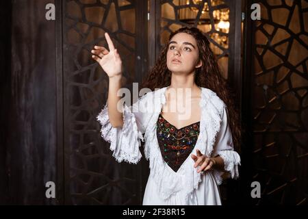 Eine junge schöne Brünette mit langen lockeren Haaren in einem Weißer Peignoir in der Art und Weise des 19th. Jahrhunderts hält Aus ihrem Hand-Cosplay für das Broadway-Musical The Phantom of Die Oper Stockfoto