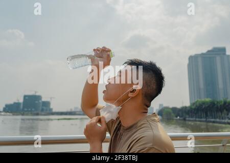 Junge asiatische Läufer Mann absetzen Gesichtsmaske, während mit Wasser in der Flasche sein Gesicht zu waschen, nachdem fertig laufen und arbeiten aus. Er trägt medizinische m Stockfoto