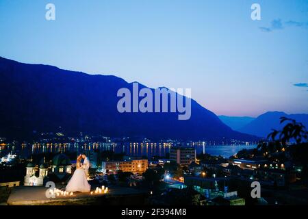 Die Braut und der Bräutigam umarmen bei Sonnenuntergang auf der Aussichtsplattform über der Altstadt von Kotor, hinter ihnen der abendliche Hafen in Lichtern Stockfoto