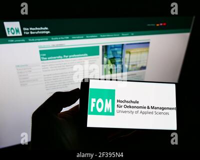 Person, die das Smartphone mit dem Logo der Deutschen FOM Hochschule auf dem Bildschirm vor der Website hält. Konzentrieren Sie sich auf die Telefonanzeige. Stockfoto