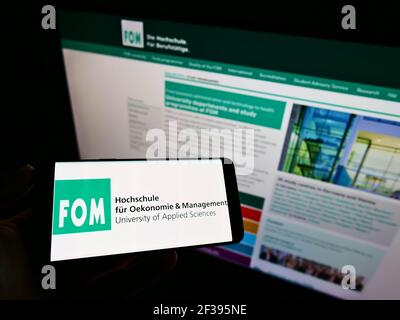 Person, die das Mobiltelefon mit dem Logo der Deutschen FOM Hochschule auf dem Bildschirm vor der Webseite hält. Konzentrieren Sie sich auf die Telefonanzeige. Stockfoto