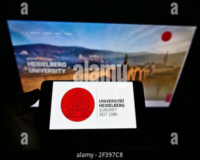 Person, die das Mobiltelefon mit dem Logo der deutschen Bildungseinrichtung der Universität Heidelberg auf dem Bildschirm vor der Webseite hält. Konzentrieren Sie sich auf die Telefonanzeige. Stockfoto