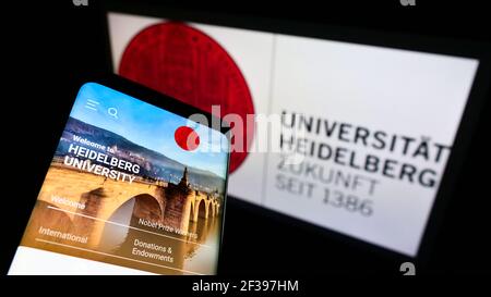 Person mit Mobiltelefon und Webseite der Deutschen Universität Heidelberg auf dem Bildschirm vor dem Logo. Konzentrieren Sie sich auf die obere linke Seite des Telefondisplays. Stockfoto
