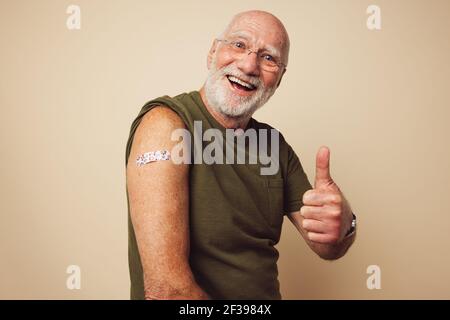 Porträt eines älteren Mannes, der lächelt und nach dem Impfen Daumen nach oben zeigt. Reifer Mann mit weißem Bart sitzt vor braunen Hintergrund Gefühl Stockfoto