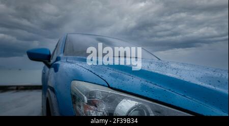 Blue Luxus SUV Auto mit Wassertropfen geparkt auf der Straße neben Meer Strand an regnerischen Tag mit Sturm Himmel. Regentropfen auf blauem SUV-Auto. Reise mit dem Auto. Elektroauto Stockfoto