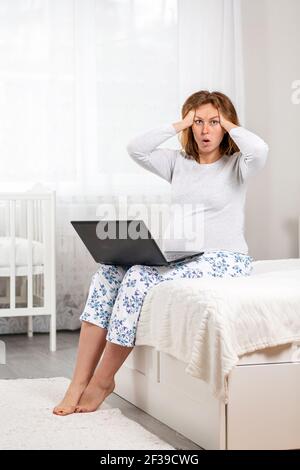 Eine schwangere Frau sitzt auf einem Bett mit einem Laptop auf ihrem Schoß und hält ihren Kopf mit ihren Händen im Schock. Weißer Raum im Hintergrund. Das Konzept von Stockfoto