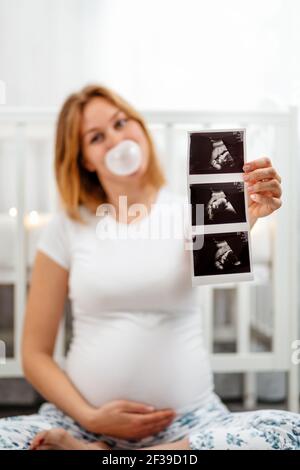 Eine junge schwangere Frau in einer Unschärfe, bläst ihr Zahnfleisch auf, umarmt ihren Bauch und hält ein Ultraschallbild in ihren Händen. Konzept der Schwangerschaft und glückliche Motte Stockfoto
