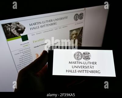 Person, die das Mobiltelefon mit dem Logo der deutschen Martin-Luther-Universität Halle-Wittenberg auf dem Bildschirm vor der Webseite hält. Konzentrieren Sie sich auf die Telefonanzeige. Stockfoto