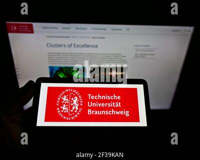 Person, die Mobiltelefon mit Logo der TU Braunschweig auf dem Bildschirm vor der Webseite hält. Konzentrieren Sie sich auf die Telefonanzeige. Stockfoto