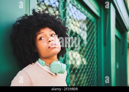 Afrohaarige Frau mit Kopfhörern, die sich an der grünen Wand lehnen. Porträt der glücklichen latina Frau . Hochwertige Fotos Stockfoto