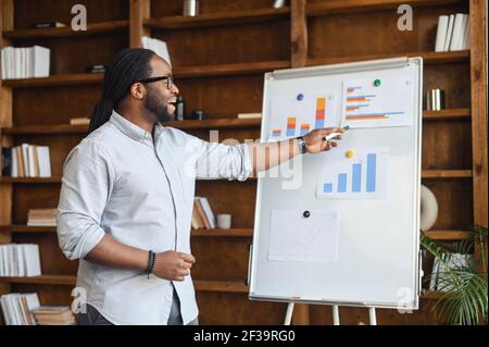 Afrikanischer männlicher Mitarbeiter mit Dreadlocks zeigt Grafiken auf einem Whiteboard, spricht über den Geschäftsplan, spricht mit einem Publikum, präsentiert Projektstrategie mit Ideen, hält Videokonferenz Stockfoto