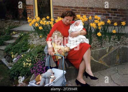 1960s sind historische, frühlingshafte und Narzissen draußen, als eine Mutter draußen in einem felsigen Garten sitzt, ihr neues Baby in ihrem linken Arm hält und die Hand ihrer jungen Tochter hält, die eine Puppe und ihren kleinen Kinderwagen mit weichen Spielsachen trägt, England, Großbritannien. Stockfoto