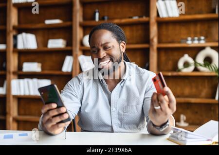 Portrait charmante afroamerikanische Geschäftsmann in Casual Shirt, hält eine Kunststoff-Kreditkarte in der Hand, bezahlen online mit Handy, Bestellungen Transaktion über das Internet, menschliche Emotionen Stockfoto