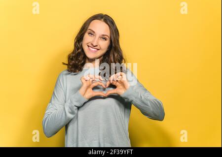 Lächelnd attraktive Brünette Frau schaut auf die Kamera und zeigt Herzform mit ihren Handflächen, gestikulende Liebe, romantische Stimmung Konzept, isoliert auf gelb Stockfoto