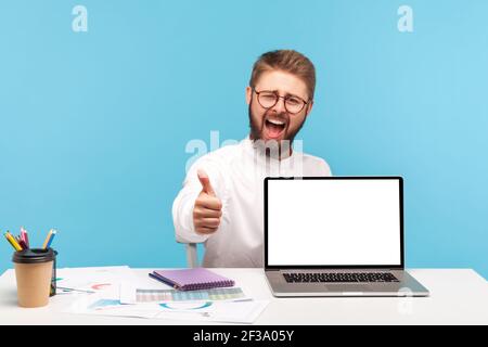 Sehr zufrieden, ein Mann, der den Daumen nach oben zeigt, sitzt am Arbeitsplatz mit leerem Bildschirm Laptop, zufrieden mit dem neuen pc, Werbebereich. Stockfoto