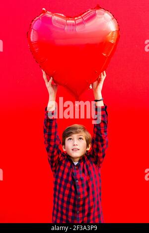 Junge hält einen großen roten Herz geformten Ballon gegen ein Rote Wand Stockfoto