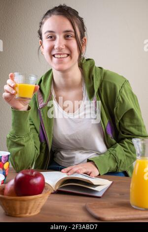 Junge Frau lächelnd mit einem Glas Saft in der Hand und einem Buch, sitzend, in Sportkleidung, hinter einem Frühstückstisch mit Saft, Äpfeln und Kaffee, Stockfoto