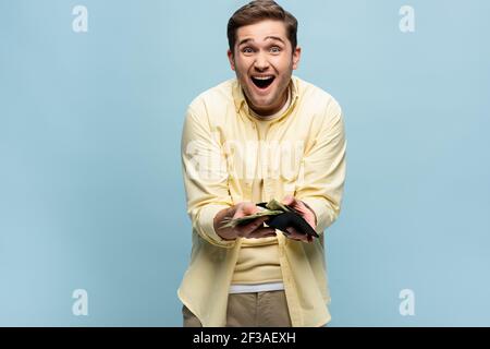 Aufgeregt Mann in gelbem Hemd hält Brieftasche mit Dollar isoliert Auf blau Stockfoto