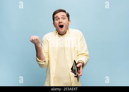 Aufgeregt junger Mann in gelbem Hemd hält Brieftasche mit Dollar Isoliert auf blau Stockfoto
