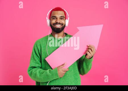 Aufgeregt junger afroamerikanischer Mann 20s in lässiger Streetwear zeigt mit Pfeil isoliert auf rosa Hintergrund, trägt Kopfhörer Stockfoto