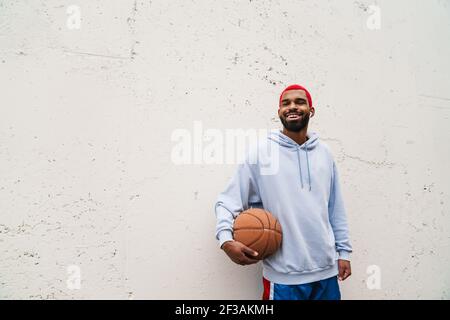 Lächelnder afroamerikanischer Kerl in Ohrhörern, der mit Basketball im Freien steht Stockfoto