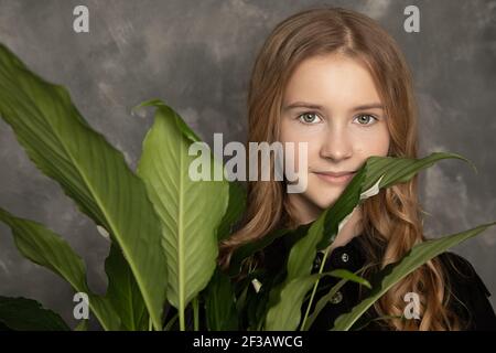 Teenager-Mädchen auf dem Hintergrund der Betonwand hält Blume mit grünen Blättern. Nahaufnahme im Hochformat. Konzept der Pflanzenpflege, Naturkosmetik für adolesce Stockfoto