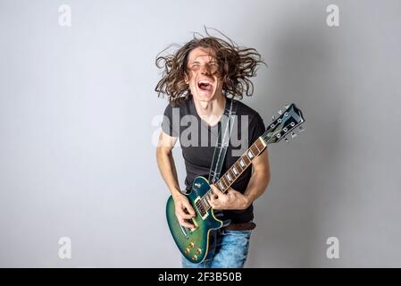 Ein junger Mann spielt eine E-Gitarre und singt und winkt mit seinen langen Haaren. Emotionale Performance von Rock and Roll Stockfoto