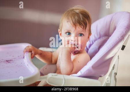 Adorable Baby essen auf Babystuhl und machen ein Durcheinander. Kleines Mädchen essen Essen in der Küche. Stockfoto