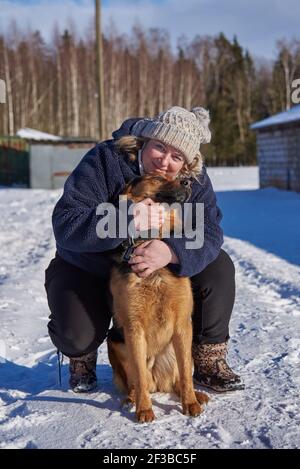 Eine dicke blonde Frau mittleren Alters in einem gestrickten Hut umarmt einen Mungel, der an einem sonnigen Wintertag auf weißem Schnee sitzt. Stockfoto