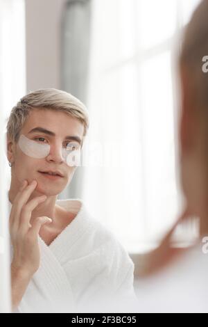 Minimal Portrait von blonden jungen Mann Anwendung Gesichtscreme und Patches, während in Spiegel während der morgendlichen Hautpflege Routine suchen, kopieren Raum Stockfoto