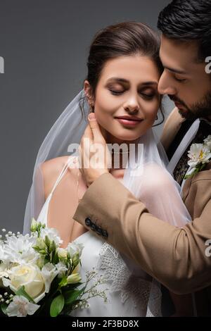 Glückliche, elegante Frau mit geschlossenen Augen in der Nähe von muslimischen Bräutigam isoliert auf grau Stockfoto