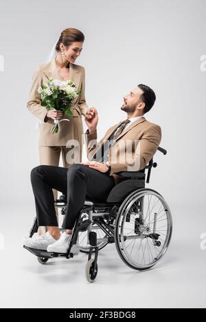 Glückliche Frau mit Hochzeit Blumenstrauß hält Hände mit behinderten araber Bräutigam auf Grau Stockfoto