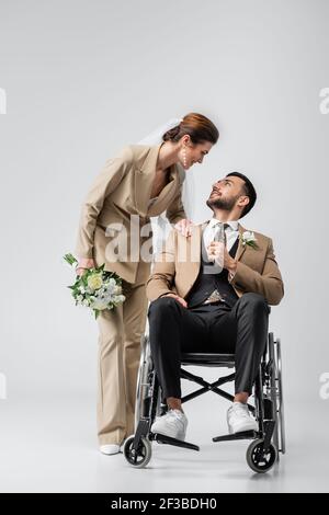 Elegante Frau mit Hochzeit Bouquet berühren Schulter von behinderten araber Bräutigam auf Grau Stockfoto