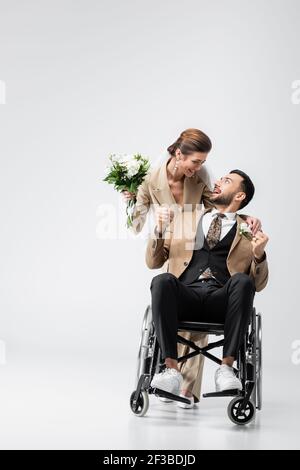 Behinderter muslimischer Verlobter, der eine Geste in der Nähe einer glücklichen Braut zeigt Hochzeitsstrauß auf grau Stockfoto