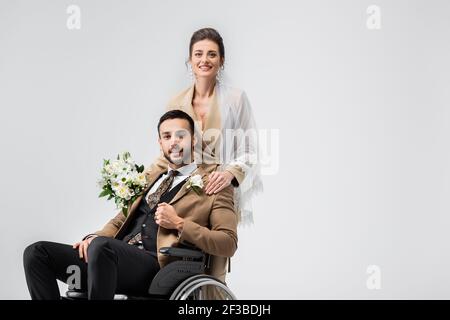 Elegante Frau im Schleier, mit Brautstrauß, Blick auf Kamera in der Nähe von behinderten muslimischen Verlobten isoliert auf grau Stockfoto