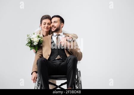 Elegante Frau mit Hochzeit Blumenstrauß hält Hände mit behinderten araber Bräutigam isoliert auf grau Stockfoto