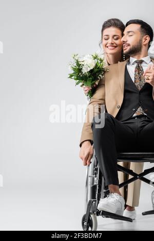 Braut mit Brautstrauß und muslimischer Mann im Rollstuhl halten Hände mit geschlossenen Augen auf grau Stockfoto