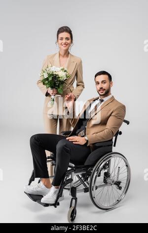 Elegante Frau mit Hochzeitsstrauß und behinderter muslimischer Mann, der die Kamera auf grau anschaut Stockfoto