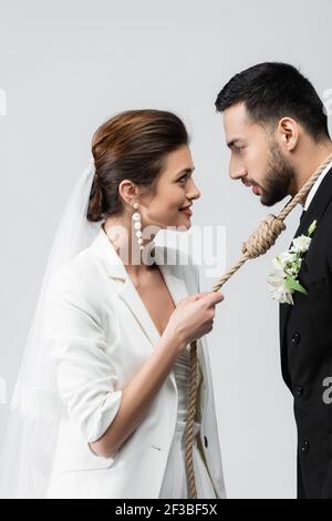 Seitenansicht der lächelnden Braut, die Seil um den Hals zieht muslimischer Bräutigam isoliert auf grau Stockfoto