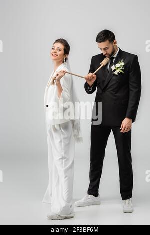 Lächelnde Braut in Hochzeitskleidung und Schleier ziehen Slip Knoten Am Hals des muslimischen Bräutigams auf weißem Hintergrund Stockfoto
