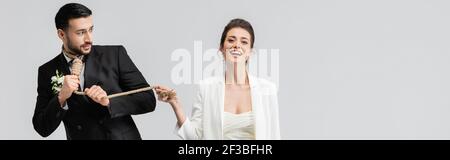 Lächelnde Braut zieht Schlupfknoten am Hals des verängstigten muslimischen Bräutigam isoliert auf grau, Banner Stockfoto