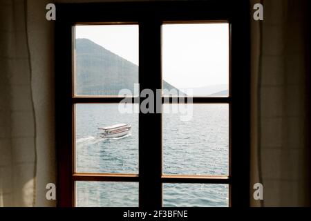 Blick vom Fenster auf die Berge der Stadt Perast und die Bucht von Kotor mit einem vorbeifahrenden Motorboot. Stockfoto